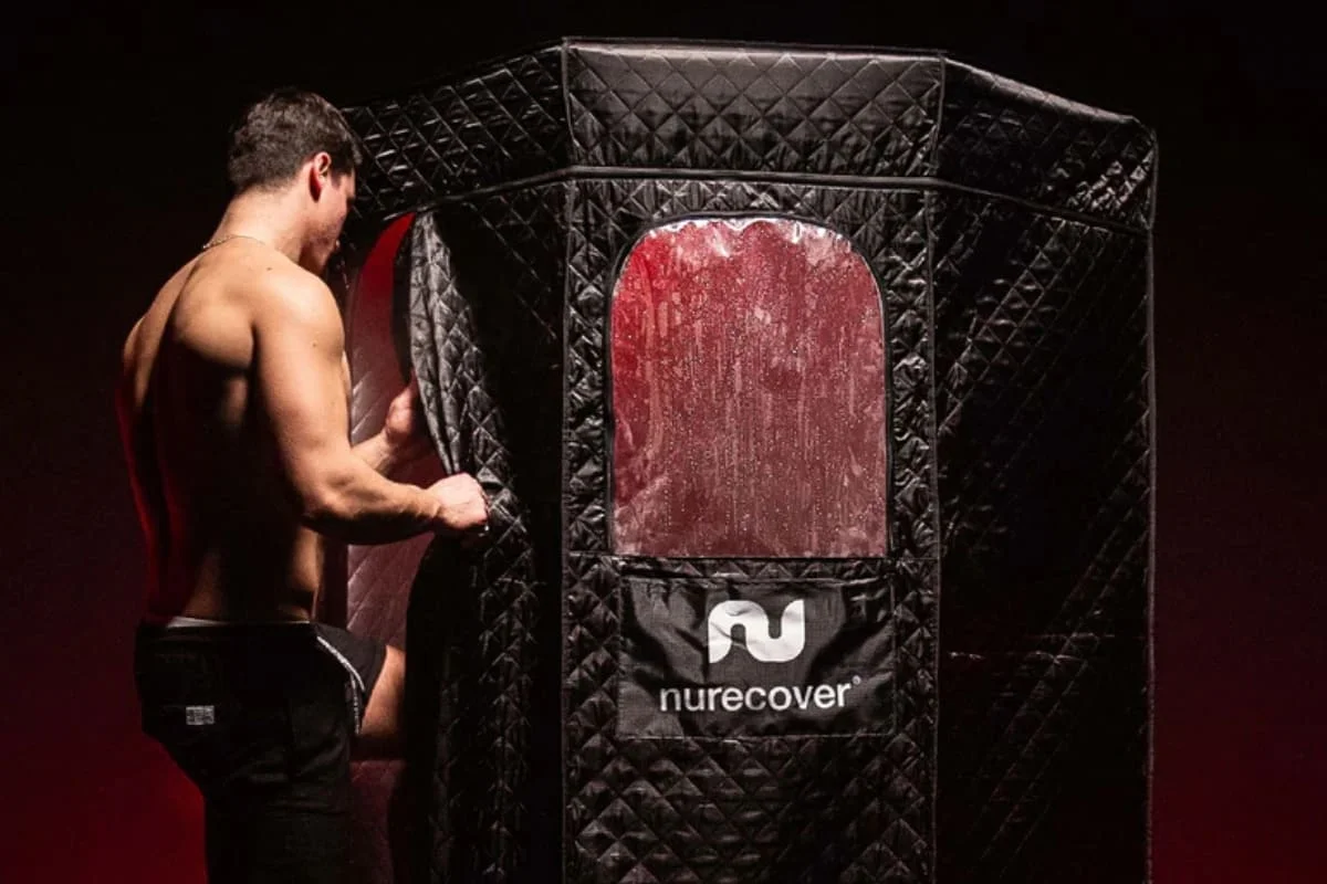 Shirtless man opening the black quilted Nurecover portable sauna showing red light therapy through transparent window panel