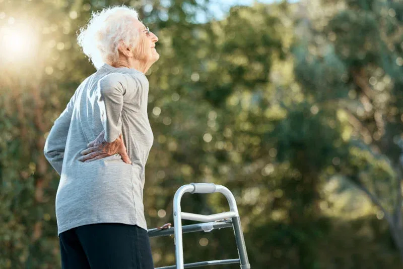 Elderly woman with white hair experiencing back pain due to osteoporosis issue, using a walking frame outdoors in a sunlit natural setting.