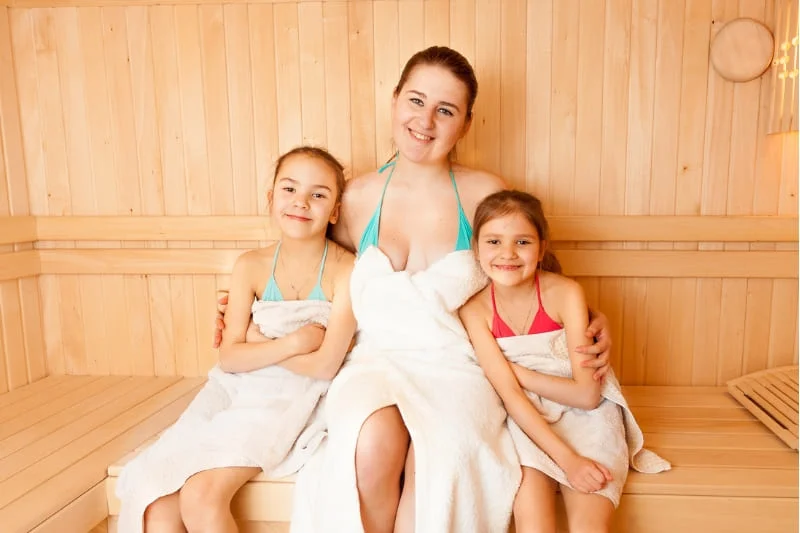 Mother and two daughters enjoying quality family time in a wooden sauna, all smiling and wrapped in white towels, demonstrating age-appropriate sauna use for children in a safe environment.