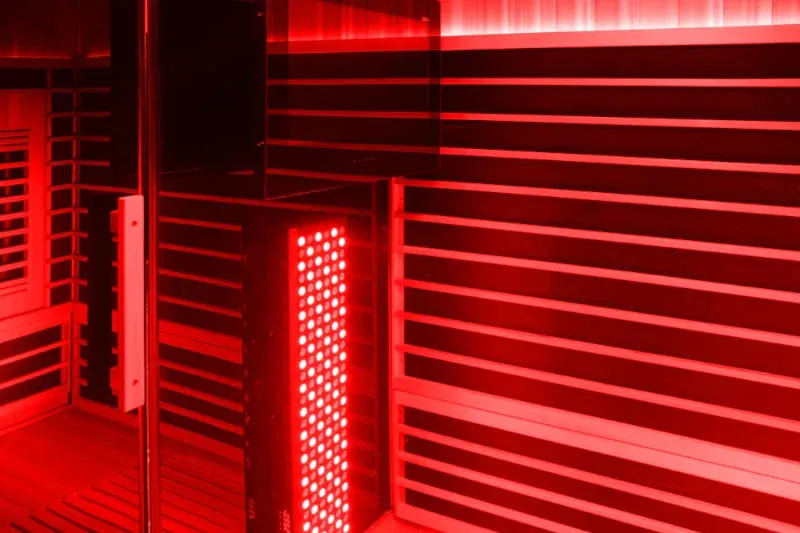 Red light therapy panel mounted on wooden sauna wall, emitting bright red light through multiple LED bulbs in a dark sauna environment.