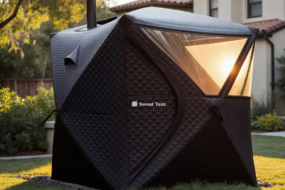 A Sweat Tent portable sauna at dusk with warm light glowing through its transparent window panel. The black quilted structure features a chimney emitting steam, positioned in a residential backyard with a house visible in the background.