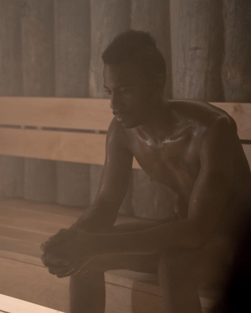 A pensive man sitting in a steam-filled sauna, with glistening skin, contemplating the potential calorie burn from the heat exposure.
