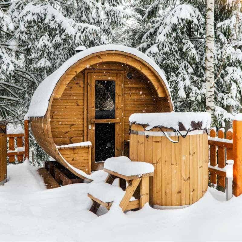 An outdoor barrel sauna covered with snow in a scenic winter forest, depicting how extreme weather conditions can limit your sauna use in case of an outdoor sauna