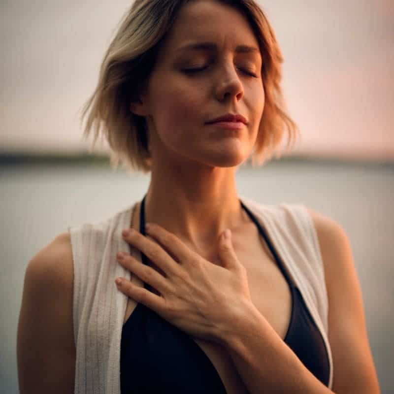 A woman in a meditative state by the water, hand on her chest, exuding calmness and well-being that mirrors the heart health benefits associated with using a sauna.