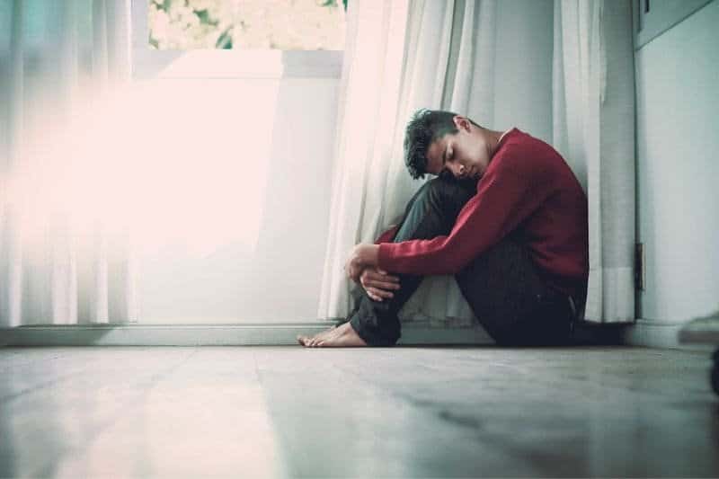 A young man seated on the floor, resting his head on his knees, with a look of contemplation, by a bright window casting a soft light around him.