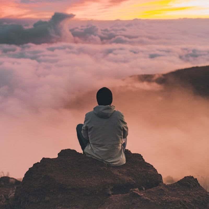 A solitary figure sits atop a mountain, enveloped by a sea of clouds during a tranquil sunset, a moment of peace that echoes the relaxation benefits of a sauna.