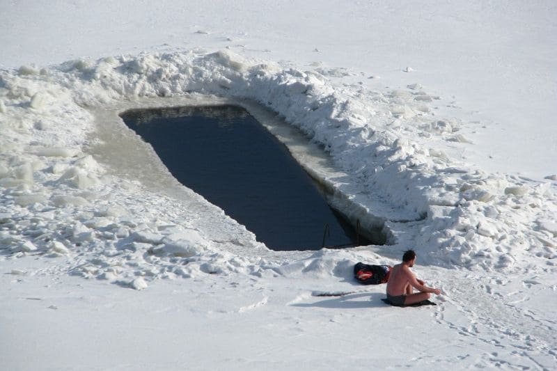 A person sitting beside a hole cut in the ice, preparing for an ice bath in a snowy landscape, demonstrating extreme cold therapy.