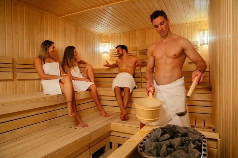 Group of people practicing sauna etiquette, with a man pouring water on hot stones to increase steam, in a communal sauna setting.