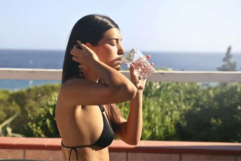 A woman in a bikini hydrates by drinking water, with a clear blue sky and the sea in the background, emphasizing the necessity of hydration during sauna sessions.