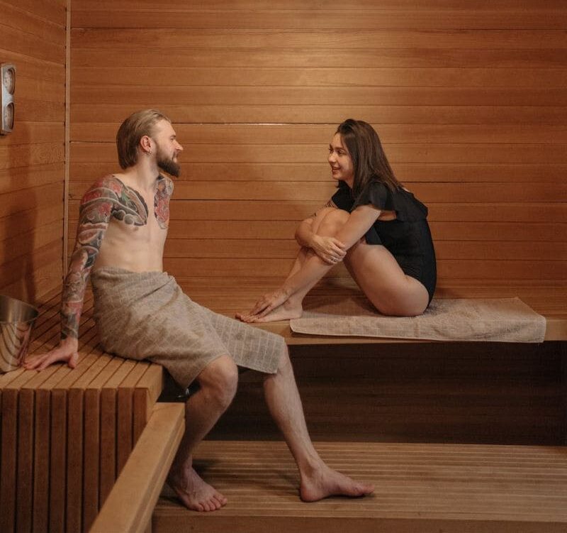 A couple in a sauna with the woman sitting correctly on a towel on a sauna bench, adhering to sauna etiquettes for hygiene and comfort.