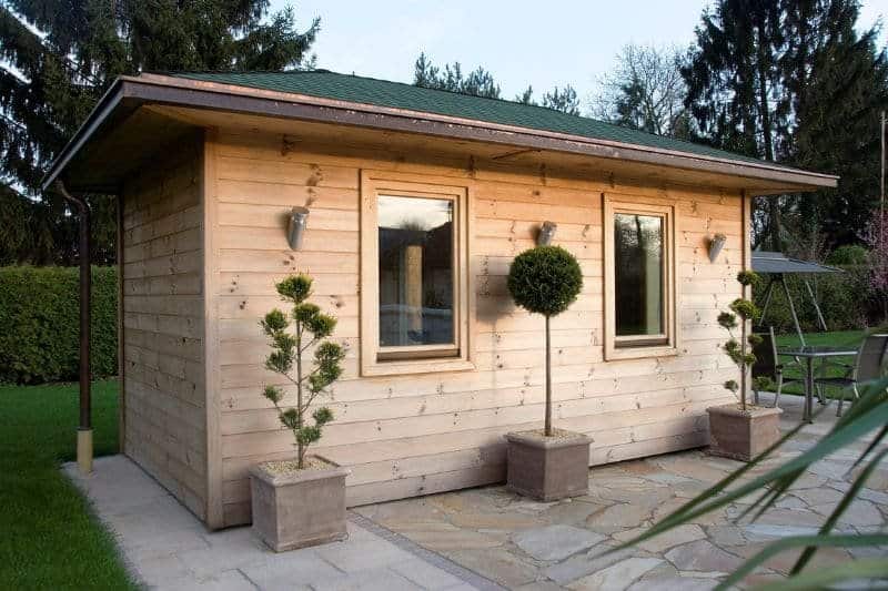 A modern garden sauna cabin with large windows and potted topiary plants, showcasing a stylish DIY outdoor sauna in a serene backyard setting.