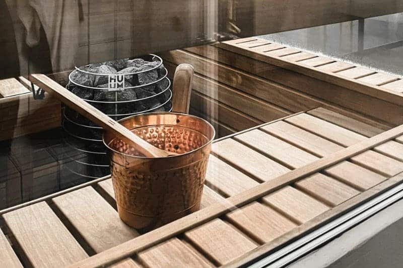 Interior of a sauna with an electric heater and a copper ladle and bucket on wooden benches.