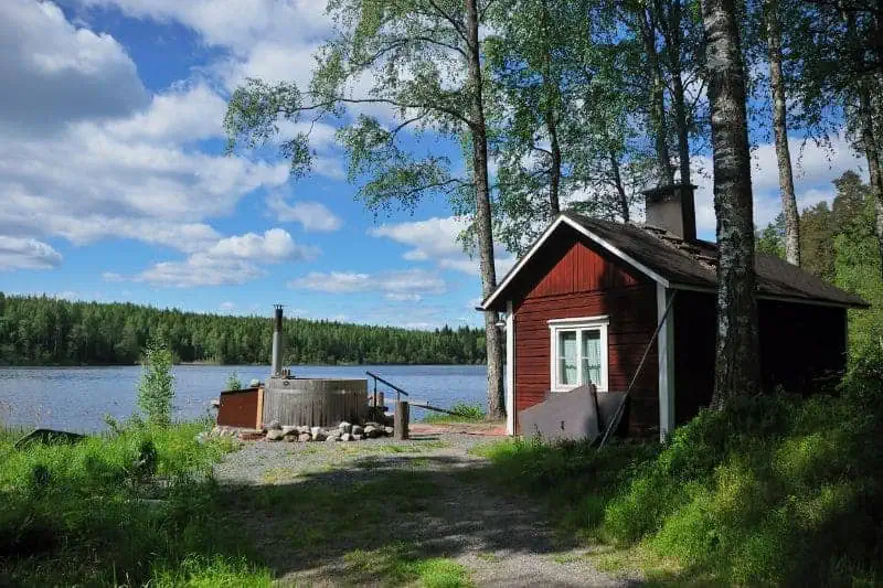 Traditional red Finnish sauna by a serene lakeside, surrounded by lush green trees under a clear blue sky, embodying the tranquil essence of Finnish sauna culture.