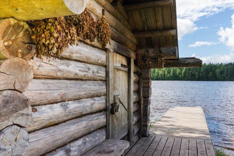 Traditional lakeside log sauna, adorned with dried birch branches, overlooking a tranquil lake, epitomizing the peaceful Finnish sauna retreat in nature.
