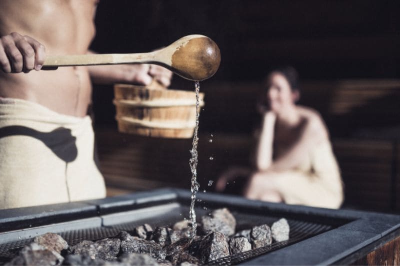 A man pouring water over hot sauna stones, with a woman enjoying the steam in the background, embodying the traditional essence of sauna bathing.