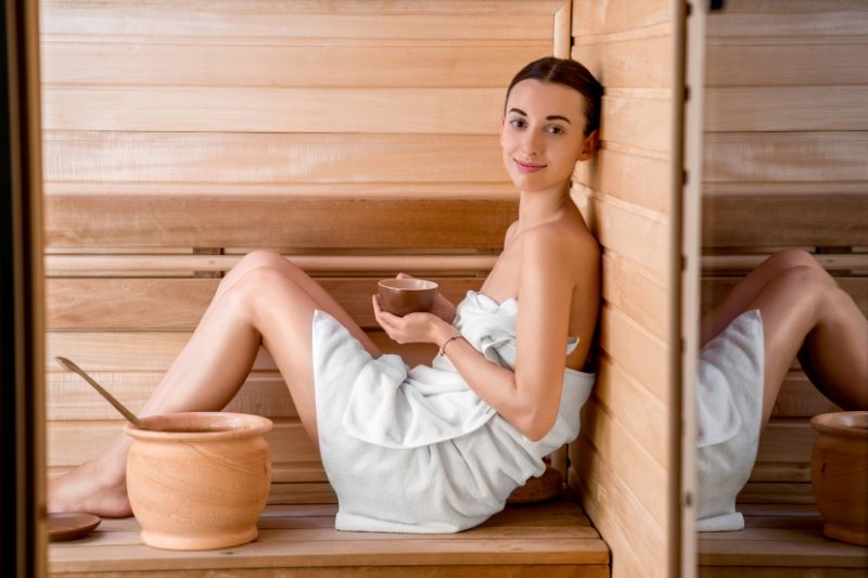 A serene young woman holding a clay cup enjoys the mental health benefits of sauna, symbolizing relaxation and wellness.