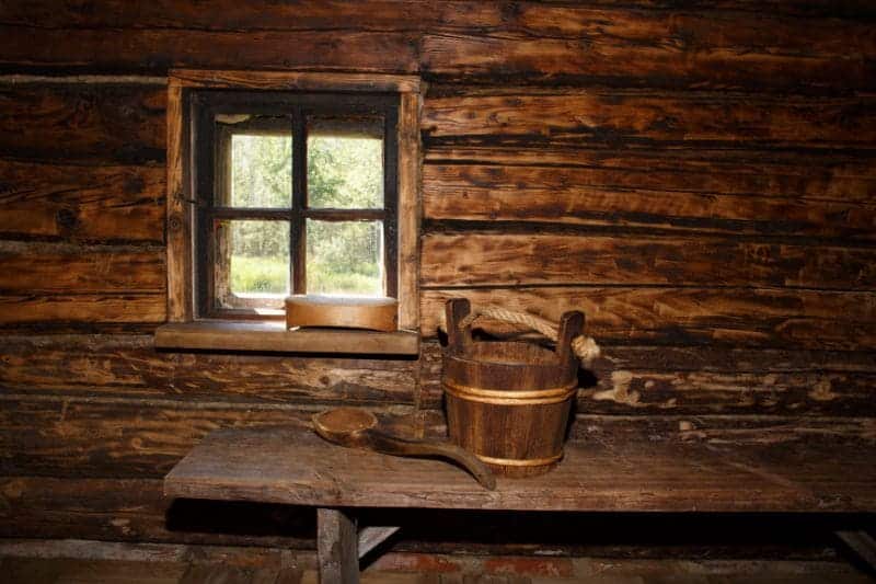 A rustic wooden bench in a traditional Finnish rural sauna, featuring a wooden bucket and ladle, with a view to the serene outdoors through an open window, evoking the authentic simplicity of the Finland sauna culture.