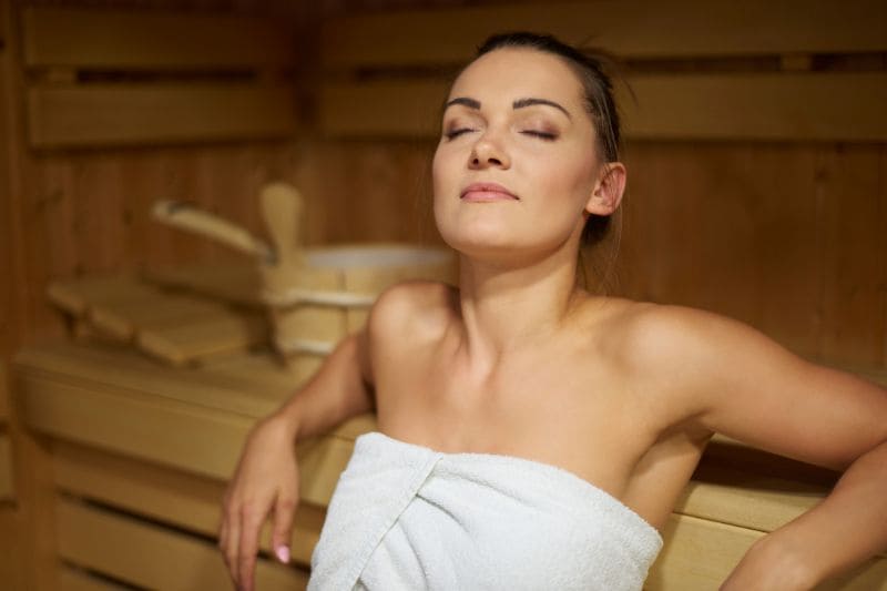 A serene woman reclines in a sauna, eyes closed in relaxation, emphasizing the tranquil and restorative aspect of sauna use.