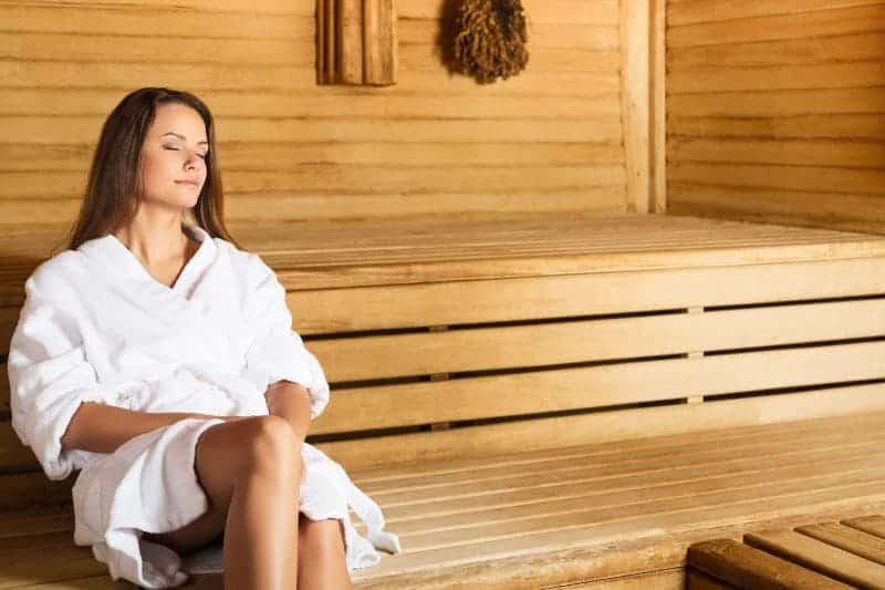 A woman in a white bathrobe with her eyes closed in a wooden sauna, trying to soak in benefits of sauna before exercise.