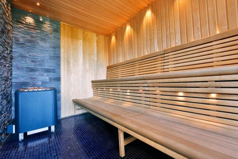 An empty, modern wooden sauna with spacious wooden benches, an electric sauna heater, and warm ambient lighting, ready for use.