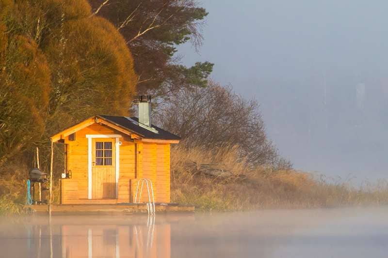 A small, charming wooden sauna by a misty lake at dawn, featuring a chimney likely connected to a Harvia stove inside. The serene setting with the sauna reflecting on the calm water surface evokes a peaceful retreat.