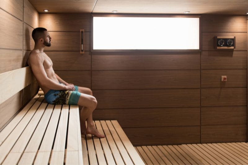 A man in colorful swim trunks relaxes in a modern wooden sauna before an intense swimming session, illustrating the benefits of sauna before workout.