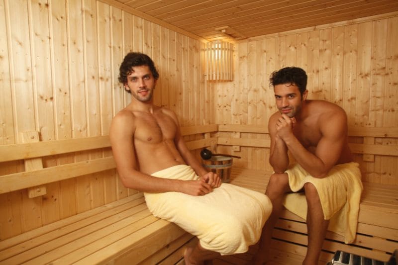Two men relaxing in a wooden sauna, sitting on bench towels around their waists, in a well-lit sauna room with wooden walls and benches.