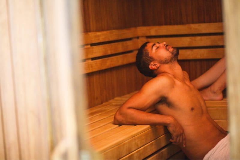 A young man in a sauna, wearing only a towel, leaning back and relaxing on a wooden bench, basking in the warmth and steam of the sauna.