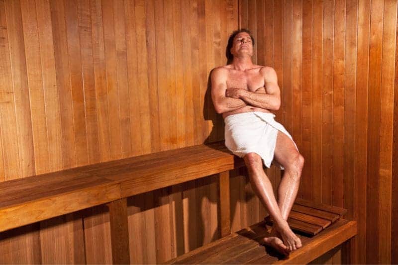 Middle-aged man sitting serenely in a sauna, arms crossed and eyes closed, in a well-lit wooden interior, exemplifying relaxation and heat therapy.
