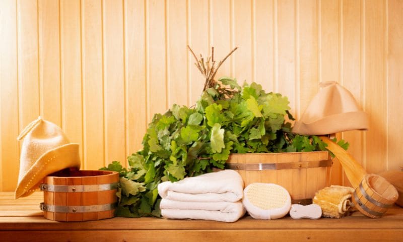 A selection of the best sauna accessories, including oak buckets, ladles, sauna hats, whisks, brushes, and towels, arranged non a wooden sauna bench.