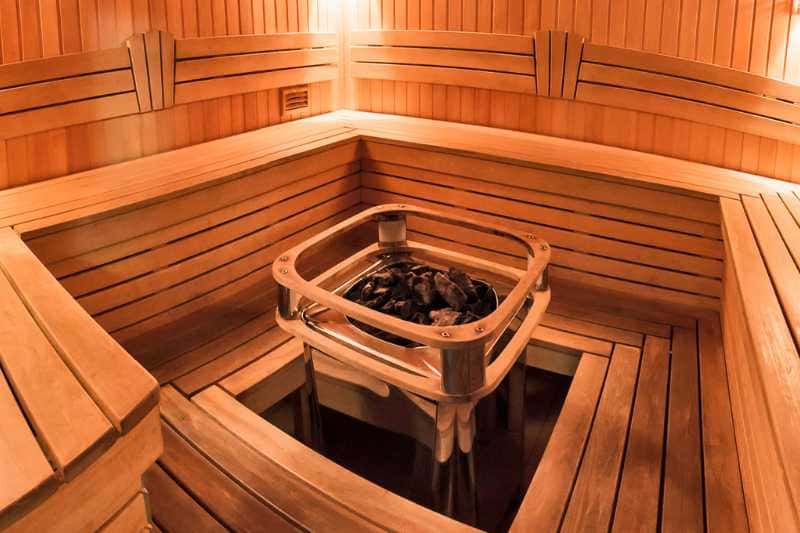 Traditional sauna heater with hot stones encased in a wooden guard at the center of a sauna room, surrounded by beautifully crafted wooden benches that enhance the warm ambiance.