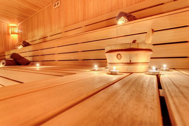 Cozy sauna setting featuring wooden benches with scattered candles providing soft light, and a traditional sauna bucket with ladle, inviting a tranquil spa experience.