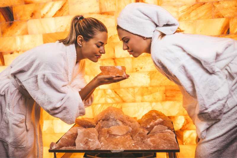 Two women in bathrobes and towels examining a large Himalayan salt crystal inside a salt sauna.