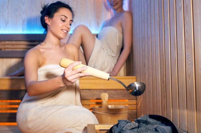 Two women sitting inside a sauna, with one of them using a ladle to pour water over the heated stones, creating steam and enhancing the sauna experience.