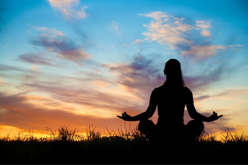 Person meditating outdoors at sunset, silhouetted against a colorful sky, embracing peace and serenity.