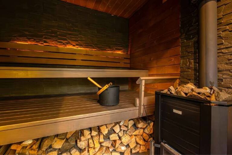 A rustic wooden sauna with a black bucket and traditional heater, logs neatly stacked underneath the bench.