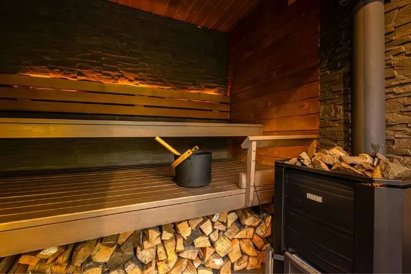 A rustic wooden sauna with a black bucket and traditional heater, logs neatly stacked underneath the bench.