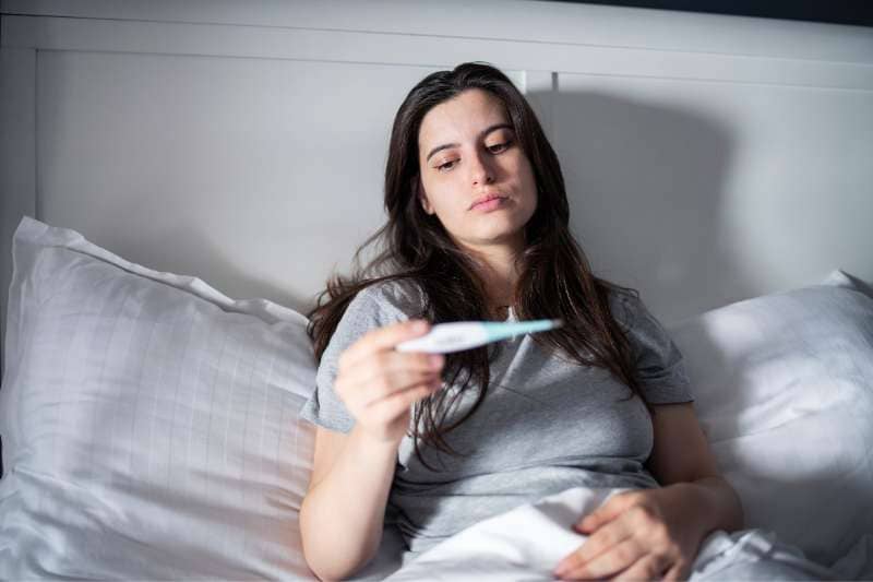 A woman in bed checking her temperature with a thermometer, looking concerned about her health.
