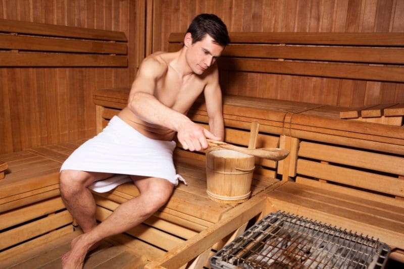 A man in a sauna, carefully pouring water from a wooden bucket onto the sauna rocks to generate steam.