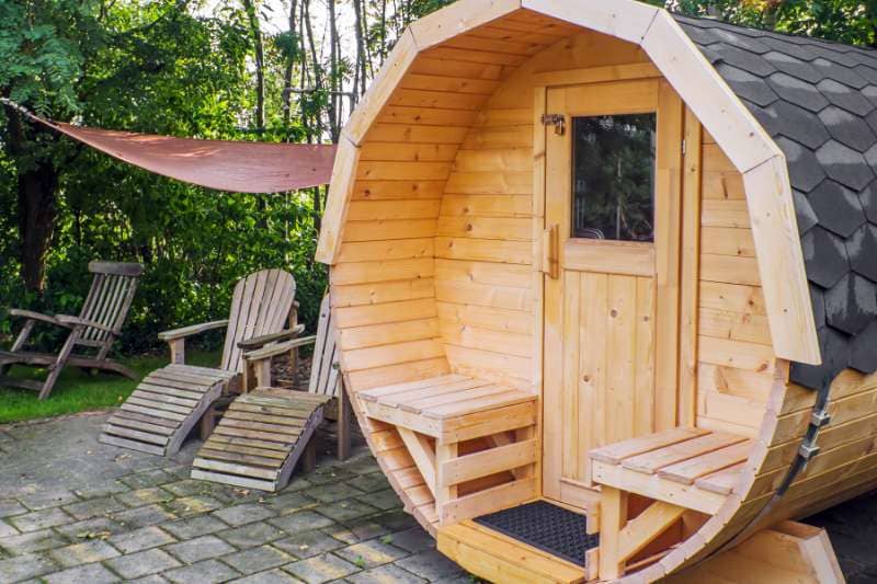 Small wooden barrel sauna with awning and patio lounge chairs on stone paved area.