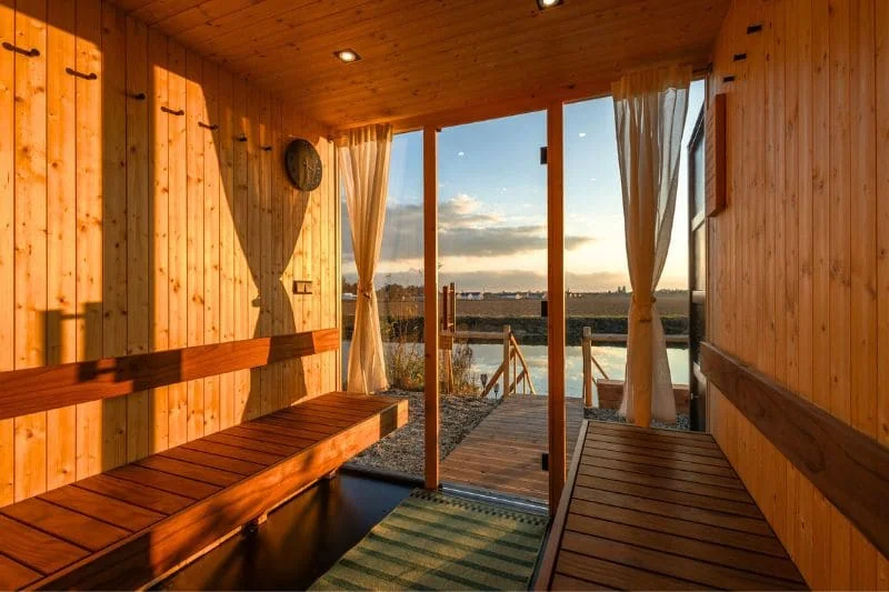 Cozy outdoor sauna with wooden benches, large windows, and a beautiful sunset view over the water.