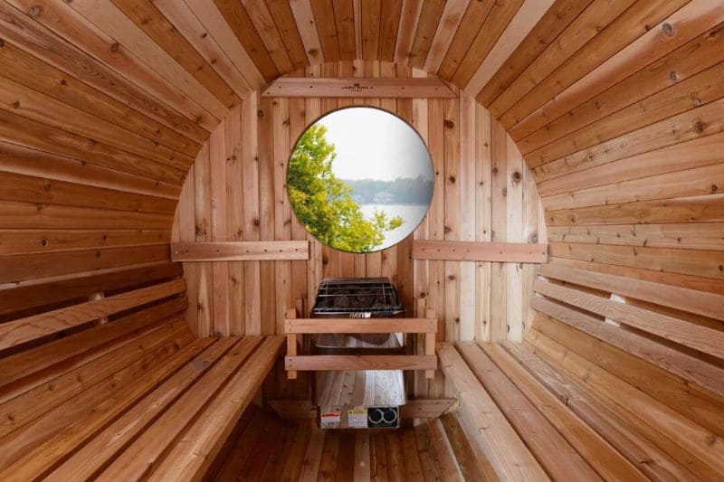 Interior of a wooden barrel sauna with a round window offering a scenic view of a lake and trees, featuring built-in benches and a heater.