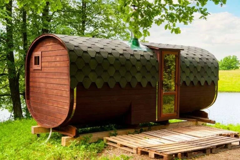 One of the best barrel sauna designs featuring a hexagonal shingled roof, situated by a serene lakeside among lush trees.