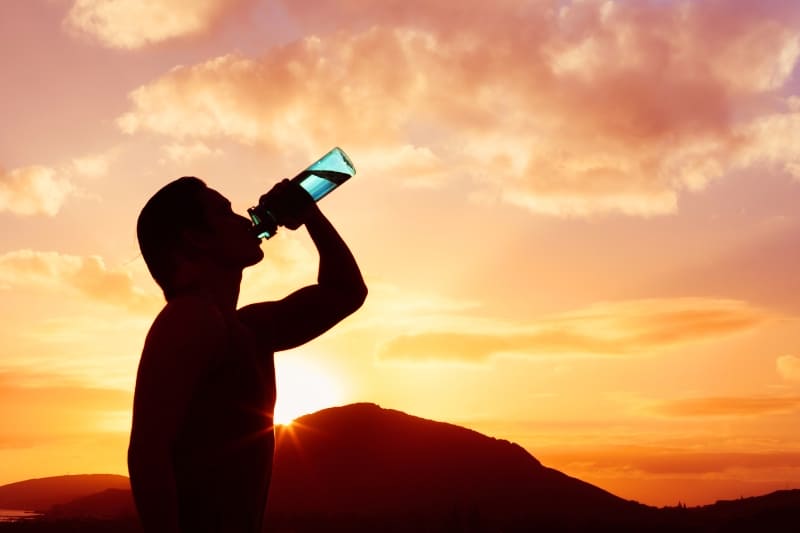 Silhouette of a man drinking water from a bottle at sunset, showcasing the importance of staying hydrated after a sauna session.