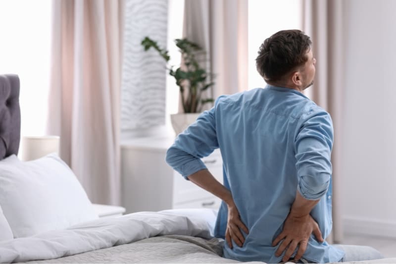 A man in a blue shirt experiencing back pain while getting out of bed in the morning.