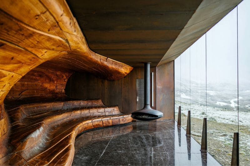 Modern wooden sauna with sculpted wooden benches and a panoramic outdoor view, featuring a unique floating fireplace.