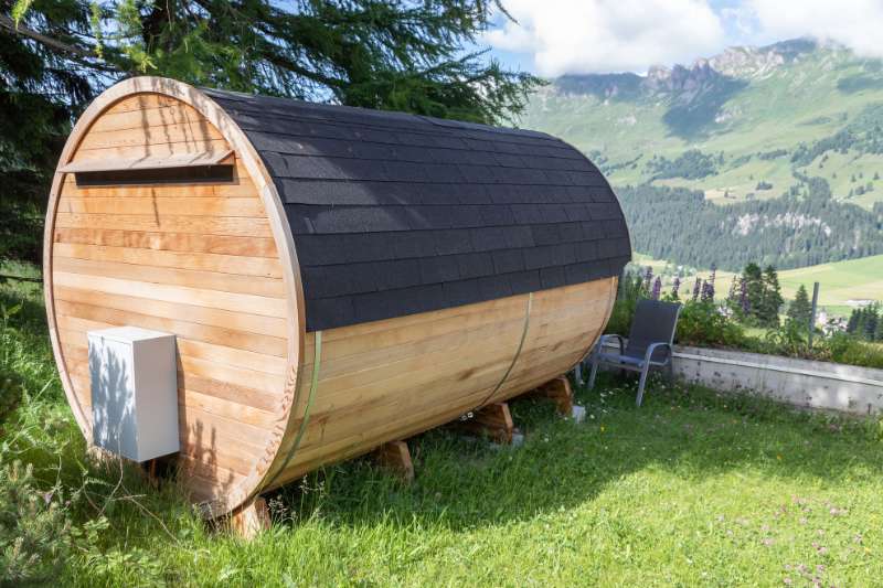 Outdoor barrel sauna with a black shingled roof, set against a backdrop of mountains and greenery, with a chair beside it.