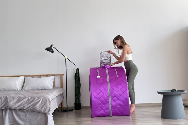 Woman setting up a purple portable sauna in a modern bedroom. The compact design makes it ideal for home use and easy storage.