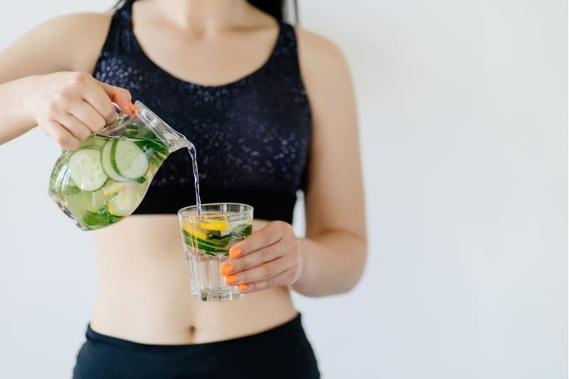 Female in a sports bra pouring cucumber and lemon-infused water into a glass, highlighting post-sauna hydration with healthy beverages.