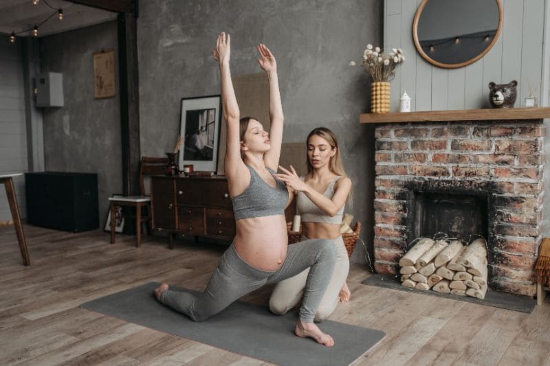 Pregnant woman practicing yoga with the guidance of an instructor in a cozy living room, enhancing flexibility and well-being during pregnancy.